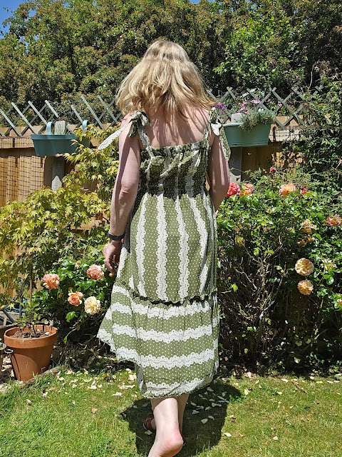 Image of woman showing back of olive green sundress in her garden. Roses in background