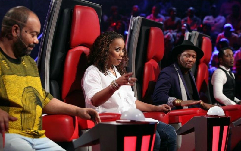Na imagem de capa: Foto do The Voice angola, mostrando as cadeiras vermelhas dos coaches de lado, todos eles negros, em destaque a única mulher que está apontando para o palco e olhando com uma expressão divertida. 