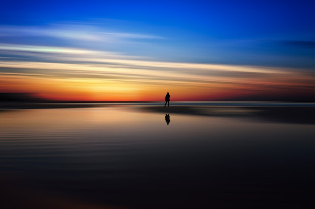 La sagoma di un uomo in piedi sulla spiaggia si staglia in lontananza contro il mare e il cielo al tramonto