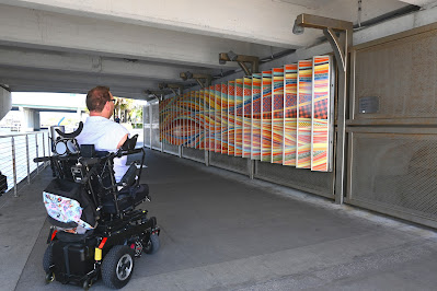 Located under the Fortune Taylor Street Bridge, is an art piece, named 'Woven Waves.' This side of the piece has a series of swirls that are orange, yellow, and green, in varying patterns. The art piece is a high contrast to the bridge around it, which is grey.