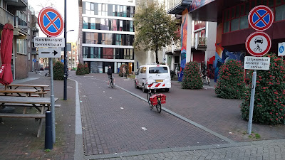 Une piste cyclable à double sens entre de grands bâtiments, avec des panneaux permettant un accès limité aux véhicules à moteur.