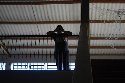 A foto foi tirada de baixo para cima, pois Marcos estava no segundo andar do ginásio. Ele está apoiado na grade de segurança, segurando a câmera nas duas mãos e apontando-a em minha direção.