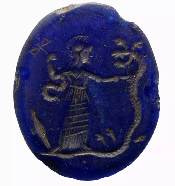 Μπλε γυάλινη πλάκα, με εγχάρακτη παράσταση θεάς με κράνος ενώ κραδαίνει ένα διπλό τσεκούρι και να πατάει ένα πολυκέφαλο φίδι. Πιθανώς από την πόλη Αχμίμ της Αιγύπτου ( 2ος-3ος αιώνας μ.Χ. )