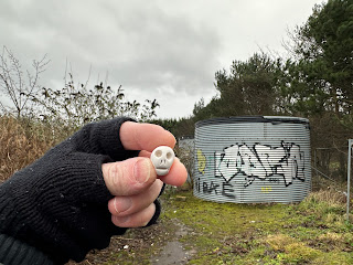 A photo of a small, ceramic skull (Skulferatu 119) being held up with a large metal container in the background.  Photo by Kevin Nosferatu for the Skulferatu Project.