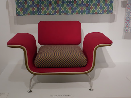 Photo of a chair by Alexander Girard. Upholstered in red, stylized design in two pieces. The back is one piece, just a flat square. The rest of the chair flows: a flat arm curves down to create the side of the chair, then the seat, then back up to make the other side, and out flat to make the other arm. There's also a large square plaid pillow on the seat.