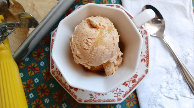 two scoops of Earl Grey Ice Cream with Lemon Curd Swirl sitting in a tea cup and saucer on a floral placemat next to the ice cream container and ice scream scoop