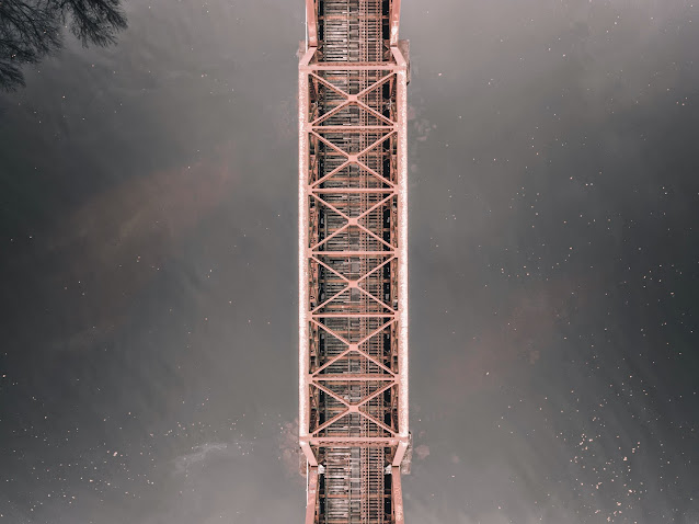 Aerial view of a long, narrow train bridge with intricate metal trusses, spanning across a calm body of water. The symmetry of the bridge's structure is highlighted by the still water below, reflecting a mirror image of its form. The overcast sky above subtly blends with the water's surface, creating an almost ethereal atmosphere around the man-made marvel.