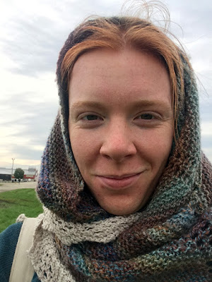 A selfie of a young, redheaded white woman with a many-colored, muted jewel-toned knitted shawl wrapped over her head and around her neck, in front of a pale, cloud-smudged sky and grassy fairground. She's smiling slightly, which makes crow's feet appear around her eyes.