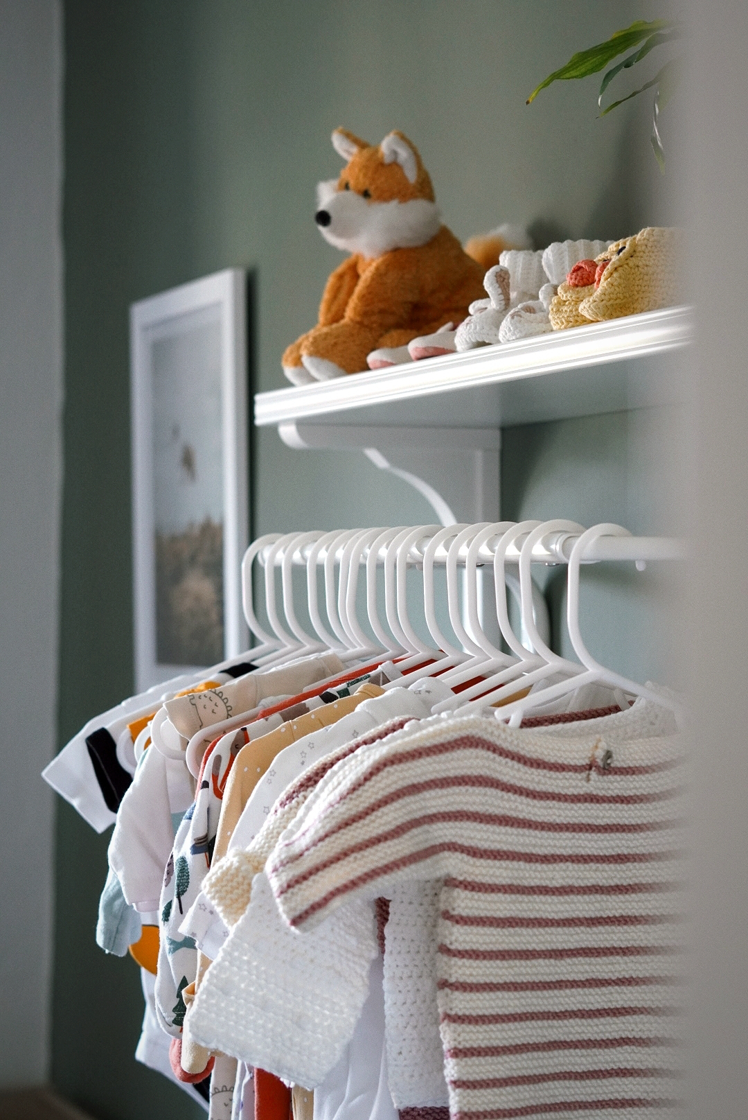 shot of a nursery; the wall is sage green and shown is a shelf, clothes rail with baby clothes hanging and a print in the background (out of focus)