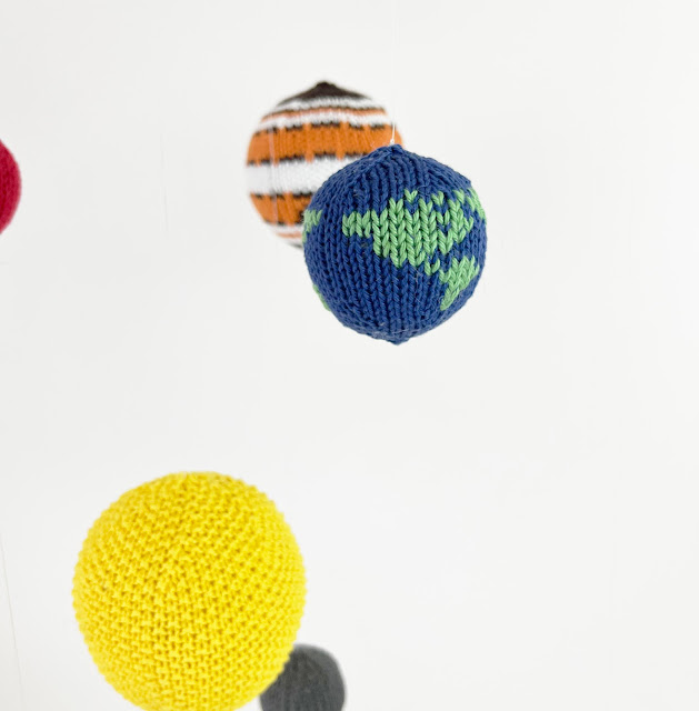 close up of knitted planet earth with sun, jupiter, mercury and mars around it as part of the solar system space mobile hanging in front of a white background