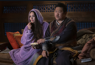 Wong and Madisynn seated on a couch. Wong is wearing his robes of off, but Madisynn is wearing a purple snuggy robe with the hood up. They're watching the screen and eating popcorn.