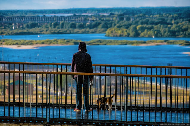 Человек с собакой на мостике на фоне реки и лесов