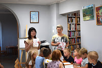 Zdjęcie przedstawia dzieci siedzące przy stolikach w czytelni Biblioteki. Maluchy biorą udział w warsztatach przeprowadzanych przez psycholog Anna Skurnicę na temat zasad dobrego wychowania.