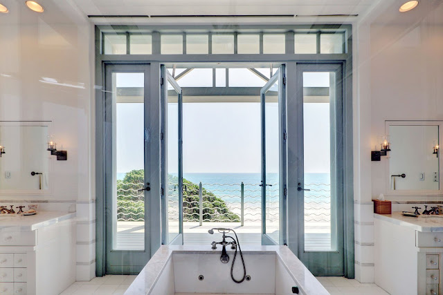 ocean view from the white master bathroom with stand alone bathtub facing blue trimmed glass doors and windows lead onto a balcony.