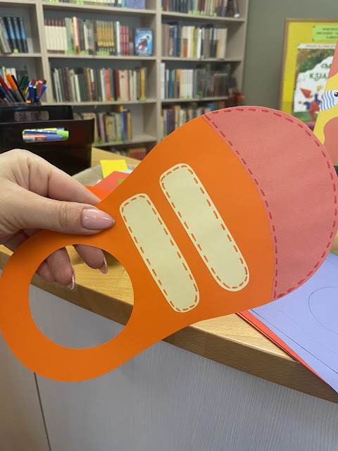 Lewa ręka trzyma papierowego, pomarańczowego buta. But ma czerwony nosek i jasnożółte rzepy. Za nią widać ladę biblioteczną. Na ladzie  widać pojemnik z flamastrami i pojemnik z klejami oraz papierowe komponenty potrzebne do wykonania pracy plastycznej. W tle widać regał z książkami.