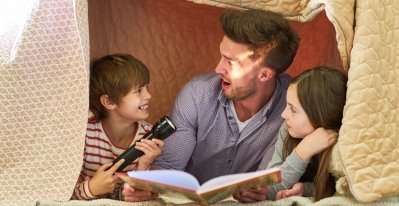 A boy and a girl are lying in a blanket fort with their arms propping them up. Their dad is reading aloud from a book, while the boy playfully shines a flashlight on his father's face.