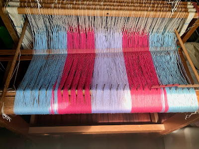 The back of a floor loom, showing a warp in the trans flag colours spread in the raddle and threaded through the heddles. There is a pair of lease sticks threaded in the warp between the warp beam and the heddles.