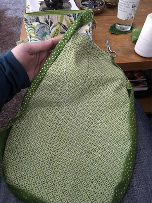 A flat-topped teardrop of green printed fabric, with gold-speckled green binding attached and partially pinned on top, and a welter of sewing notions on the low table in front of the photographer.