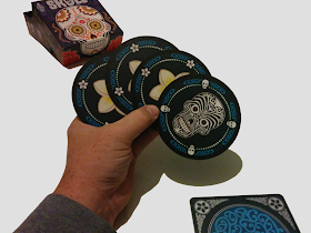 The four blue cards fanned out in a player's hand. The box, with it's Mexican folk art style artwork, is visible in the background. The design of the score mat is partially visible. The skull card, on the right in the player's hand, is covered with black lines and swirls. The flower cards are partially obscured, but you can see enough to tell that it's a white five-petaled flower.