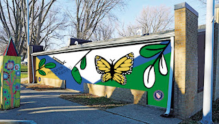 A photoshopped image of a building side, with a mural design on top of it in flat colors. The mural is white, navy blue, green, black, and yellow ochre, and shows different stages of the monarch butterfly life cycle.
