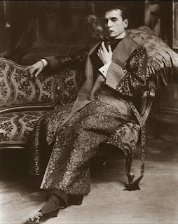 Actor William Gillette sits pensively, as 'Sherlock Holmes' in a stage production. He is smoking a pipe while seated and wearing a Victorian Housecoat which has the appearance of a long and heavy dressing gown with quilted silk lapels.