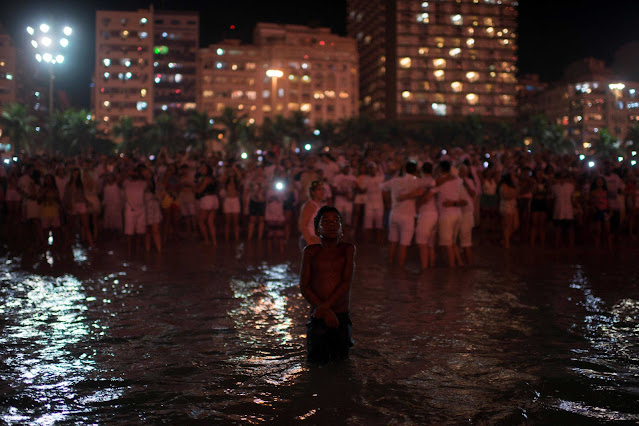 Ao centro da imagem, um menino negro sem camisa e vestindo uma bermuda. Ele está molhado e se encontra olhando para cima com a boca entreaberta e com as mãos cruzadas à frente de seu corpo. O menino está localizado à beira mar com água até os joelhos. Um pouco mais atrás, na areia da praia, está uma multidão de pessoas vestidas de branco. Alguns dão as costas ao menino para registrarem o momento em fotografias e outros olham para cima observando o céu. Ao fundo da imagem observam-se prédios bem iluminados, postes de luzes e algumas árvores. A fotografia está em cores. Tirada por Lucas Landau. Propriedade da Reuters.