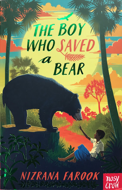 Central characters Karadi the bear, and the boy Nuwan sit within the jungle edge canopy. Karadi stand all feet on a tree stump while Nuwan layys hand up to wards Karadi's face. The title sits within a pale yellow setting sun and in a nice metalic green and pale red large font. The sky is a mix of orange red yellow and light blue. Nuwan is in blue knee length shorts and a white top. The foliage depicted around the authors named is in metalic green.