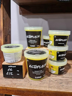 A row of large plastic clear tubs filled with bright neon yellow-green slime with a black label with ectoplasm shower slime lush in white font on a large light brown wooden shelf on a bright background