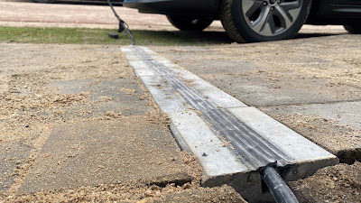 A close up view across a footway from a front garden to the wheels of a car in the road. There is a concrete channel with a rubber strip running through the middle into which a cable has been pushed. The rubber strip leaves a level footway over the cable.