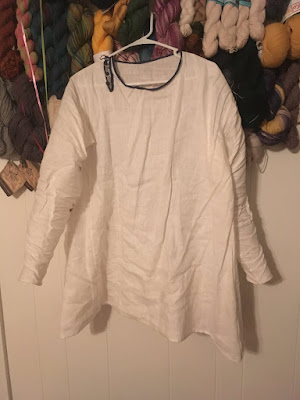 A white long-sleeved tunic with blue neck binding and buttons on a hanger, hooked on a wall in front of dozens of hanks of yarn.