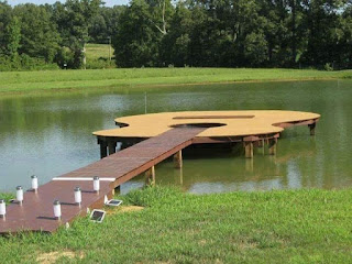 Foto em dia ensolarado. No lago do rancho Macota em Minas Gerais, circundado por um gramado bem aparado, um deck em formato de um violão gigante. Na beira do lago, assentada sobre o gramado, está a mão do violão; seis luminárias tubulares, três de cada lado, representam as tarraxas. O braço do violão, é uma ponte de acesso ao corpo do violão, onde o tampo serve de palco quando há show no rancho. Sobre a superfície do lago, o reflexo da vegetação e do deck. Ao longe, um bosque.