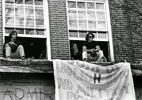 A photograph of students perched in open windows from which a hand-made banner hangs. 