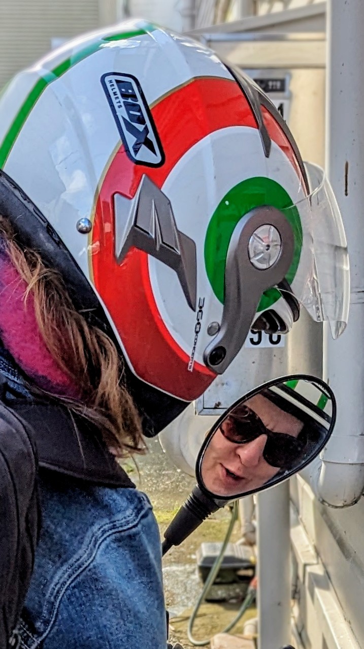 A woman from behind, sitting on her motorbike. Her face, from within a helmet, is framed perfectly in the mirror.