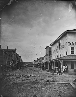 Black and white photo of street scene around 1865 with dirt street and two story buildings lining either side. People are standing in front of building to the right and a man sits on a horse on the left side of the street