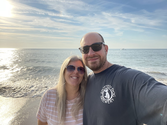 A selfie of me and Dad on another beach walk. It is before noon as the sun is still between the eastern horizon and the middle of the sky. The open ocean is behind us. I am wearing a pink and white striped t-shirt and sunglasses. My hair is straightened and is falling over my shoulders. Dad is in a black brewery tshirt (i do not know which one) and is wearing his sunglasses. We are both smiling for the camera.