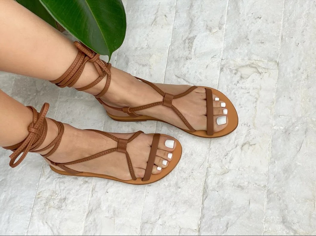 A pair of low heeled lace-up sandals in brown leather, crossed in the middle and tied at the ankle with laces. The model wears white nail polish. The background is a gray stone-effect tile floor and a ficus elastica leaf can be glimpsed