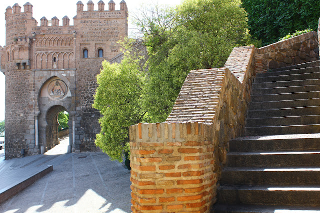 Średniowieczna brama Puerta del Sol w Toledo. Zwieńczona krenelażami ceglana brama miejska o podkowiastych łukach, wzniesiona w stylu mudejar. Na pierwszym planie schody o ceglanych barierkach, prowadzące ku kolejnej z bram miejskich.