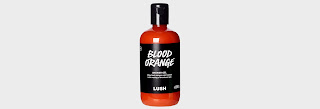 A tall cylindrical plastic bottle filled with bright orange shower gel with a black rectangular table with blood orange shower gel lush in white font with a black cylindrical lid on a bright background