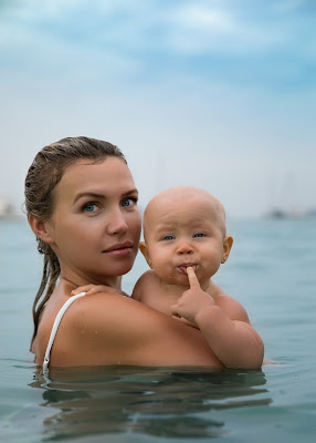 Picture of a mother looking at the camera and holding her baby whilst standing in the water. Benefits of Baby, Infant and Toddler Swimming