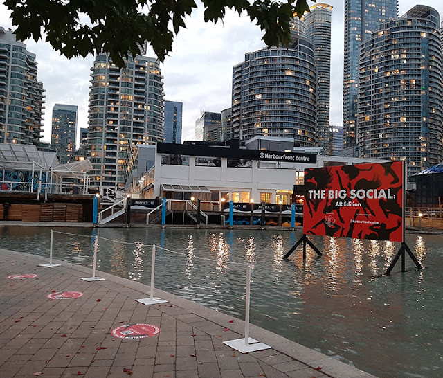 Billboard-style screen in the pond at Harbourfront. Round red circles on the pavement around the pond. Residential towers in the background.