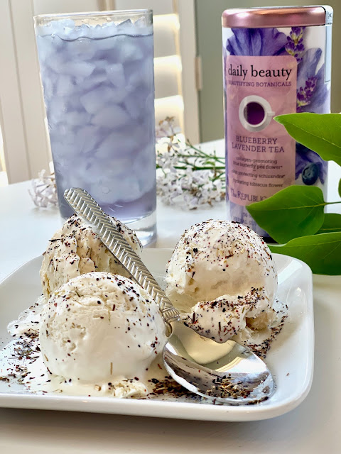 A dish of ice cream sitting on a white plate, with a glass of tea behind it and a canister of Blueberry Lavender Tea. Lilacs on the table.