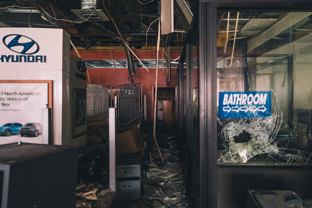 The shattered glass of a door leading to a bathroom in a derelict car dealership, amidst the chaos of abandoned promotional stands and office equipment.
