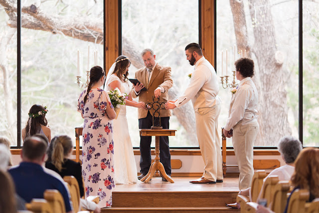 Summer and David decided on a small, intimate wedding, tying-the-knot at the tucked-away and woodsy Thunderbird Chapel in Norman, OK.  Little did Summer know when David became her personal trainer, that they would one day be married.  This was a second marriage for both, and Summer’s bridesmaid and maid of honor, and David’s best man were their respective children.    Summer was stunning in her trumpet style lace wedding dress, “killer” glittery heels, holding a beautiful, handmade bouquet consisting of pink roses, white lilies, pink freesia, white baby’s breath and greenery.  And David looked dashing in his champagne-colored, three-piece suit, light blue tie, and a single rose boutonniere. Such a beautiful couple on their wedding day!        Summer’s dad was brought to tears during his “First Look;” it was clear that this was a special day for him as well.  The littlest girls led the way down the aisle, dropping rose petals, and racing each other to the end, while David and his son watched with awe as Summer, his soon-to-be-bride, walked down the aisle, arm-in-arm with her dad.  The ceremony included the assembly of a Unity Cross, signifying the two becoming one together with their faith in Christ, and hand-written vows.  At the end of the ceremony, David, caught up in the emotion of the “I do’s,” kicked up his heel in celebration!     After the ceremony, the bride and groom were greeted by a throng of well- wishing guests blowing bubbles.  The day was filled with heartfelt moments shared by the people they loved, and we tried our best to capture every beautiful detail, big and small.