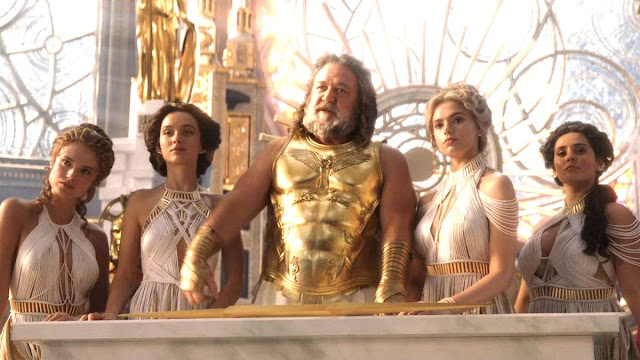 screenshot from T:L&T - Zeus (white man with shoulder length hair in a gold tunic) with two women on either side of him, all with complex braids and white halter top dresses