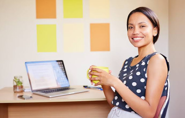 Mulher no escritório, sorrindo em frente ao notebook com uma xícara na mão.