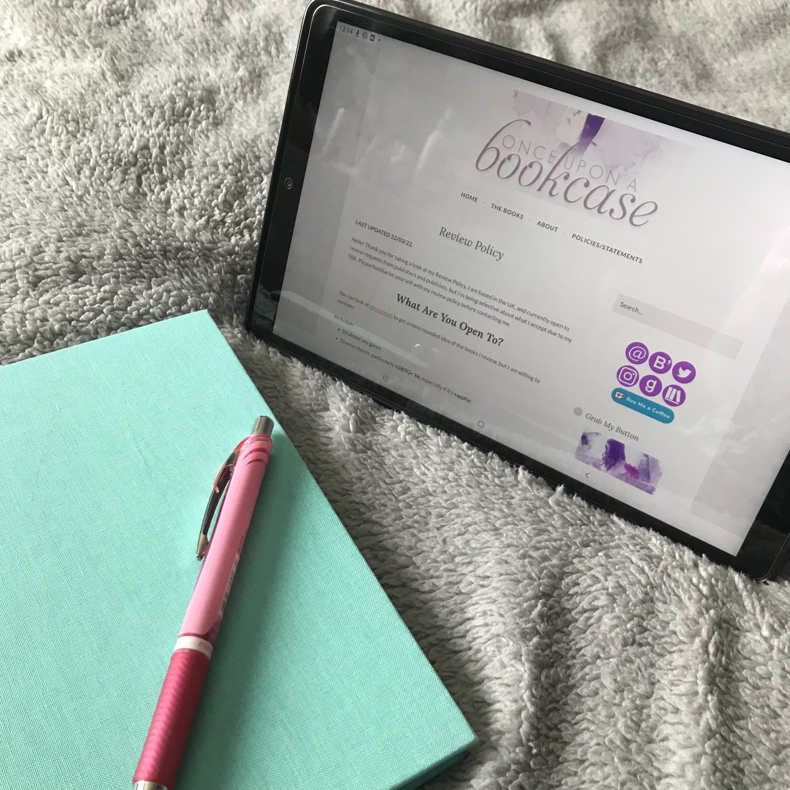 A photo of a tablet, proped up on a light grey, fluffy duvet, at an angle so the screen faces the botyom left of the photo. It's open to Once Upon a Bookcase's review policy. In front of the tablet, to the left of the photo, is a pale green, hardback notebook, and a pink pen.