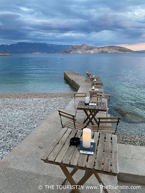 Six empty wooden tables and chairs decorated with candles on a pier over a pebbled beach and into the sea at twilight.