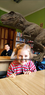 tło: sala biblioteczna. Dzieci siedzą przy stolikach. Na pierwszym planie uśmiechnięta dziewczynka obok niej wygenerowany ze smartfona obrazek dinozaura tyranozaura. W tle regały z książkami.