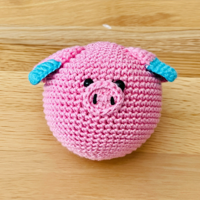 A blue and pink crocheted pig face with ears placed on a wooden table.