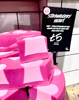 A zoomed in photo of a stack of cube shaped pieces of red soap with a white and pink heart in the centre of each slice on a light brown rectangular shelf next to a black rectangular card that says strawberry heart soap lush in white font on a bright background